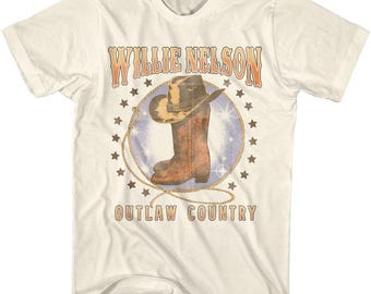 May include: A white t-shirt with a vintage-style graphic of Willie Nelson's name and a cowboy boot with a star-studded background. The text "Outlaw Country" is printed below the graphic.