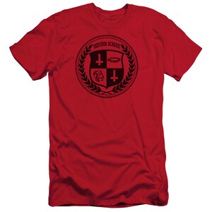 May include: Red t-shirt with a black and white crest featuring a shield with a cross, a halo, and a book. The crest is surrounded by a laurel wreath and the text "DEFORM SCHOOL".