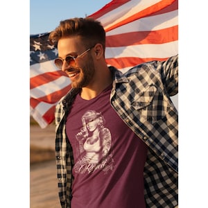 Puede incluir: Un hombre con una camiseta granate con un gráfico blanco de una mujer sosteniendo un arma y la palabra "Respeto" impresa debajo. Lleva una camisa de cuadros sobre la camiseta y gafas de sol. Está sosteniendo una bandera estadounidense detrás de él.