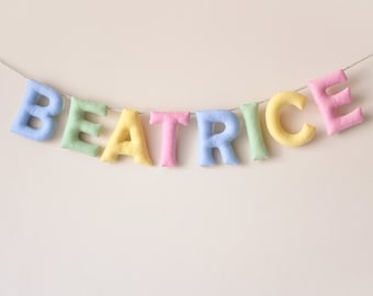 May include: A pastel-colored felt banner spelling out the name "Beatrice". The letters are strung together with white thread and hang against a white background.