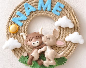 May include: A brown and tan felt animal wreath with a blue felt name "NAME" on a rope base. The wreath features a brown and white cow and a tan kangaroo.
