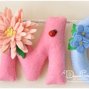 Peut inclure: Une banni&egrave;re en feutre avec les lettres "M" et "O" en rose et un "C" bleu d&eacute;cor&eacute; d'une fleur rose, une coccinelle et des fleurs bleues. La banni&egrave;re est destin&eacute;e &agrave; une chambre d'enfant ou une cr&egrave;che.