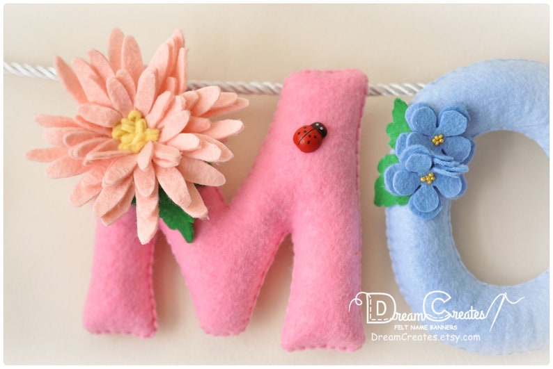 Peut inclure: Une banni&egrave;re en feutre avec les lettres "M" et "O" en rose et un "C" bleu d&eacute;cor&eacute; d'une fleur rose, une coccinelle et des fleurs bleues. La banni&egrave;re est destin&eacute;e &agrave; une chambre d'enfant ou une cr&egrave;che.