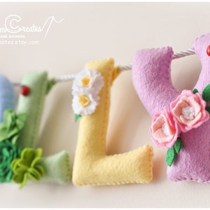 Peut inclure: Une banni&egrave;re en feutre avec le nom "Lilly" &eacute;crit en lettres color&eacute;es. Chaque lettre est d&eacute;cor&eacute;e de fleurs et de feuilles. Les lettres sont enfil&eacute;es sur une ficelle blanche.