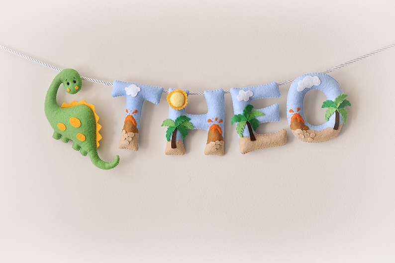 May include: A felt banner with the name 'THEO' spelled out in light blue letters. Each letter has a small volcano, palm tree, and cloud design. A green dinosaur is attached to the left side of the banner.