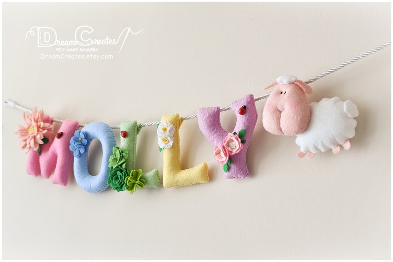 Peut inclure: Une banni&egrave;re en feutre avec le nom "MOLLY" &eacute;crit en lettres color&eacute;es. Chaque lettre est d&eacute;cor&eacute;e de fleurs et d'autres embellissements. Un mouton rose avec un corps blanc est attach&eacute; &agrave; la fin de la banni&egrave;re.