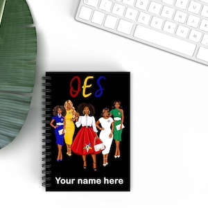 May include: Black spiral-bound notebook featuring a colorful illustration of five women in dresses. The word "OES" is written in red, yellow, and blue above the illustration. The text "Your name here" is at the bottom.