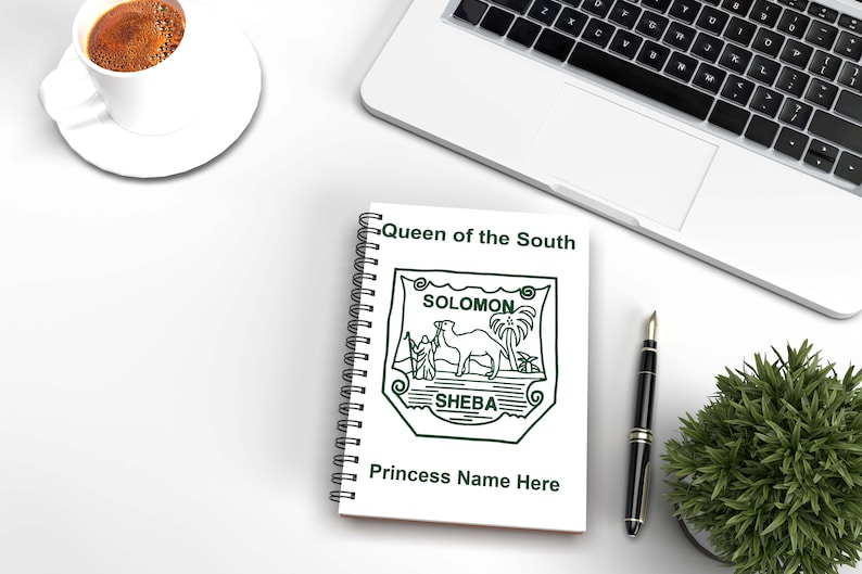 May include: A spiral-bound notebook with the text "Queen of the South" and a green emblem featuring a camel, palm trees, and the words "Solomon" and "Sheba." A laptop, pen, coffee cup, and small plant are also on the white surface.