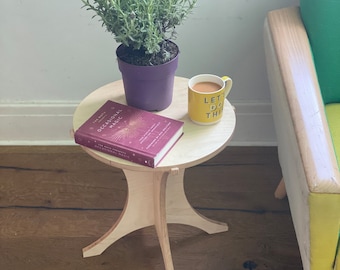 May include: A small wooden table with a round top and three legs. A book titled "The Book of Occasional Magic" is on the table, along with a purple potted plant and a yellow mug that says "Let's Do This".