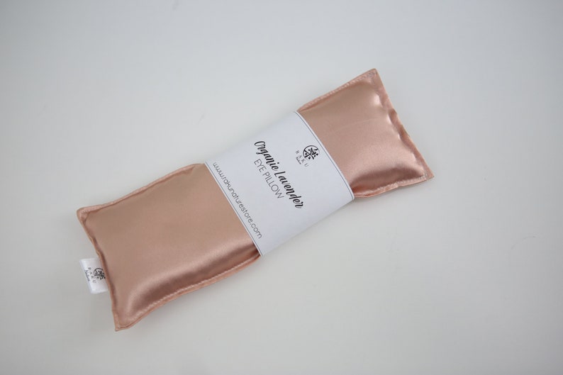 Puede incluir: Una almohada para los ojos de color rosa dorado con una etiqueta blanca que dice "Organic Lavender Eye Pillow" y "www.naturesrestore.com".