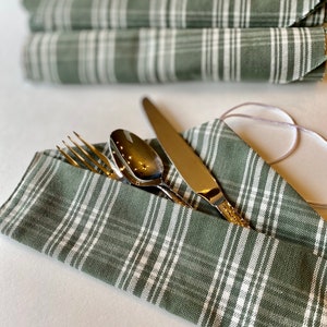 May include: A set of sage green and white plaid fabric cutlery pouches containing a silver-toned fork, spoon, and knife. The pouches feature a folded design with a thin white tie, and a second rolled pouch is visible in the blurred background.