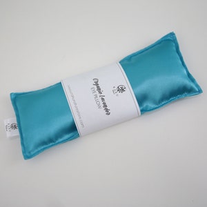 Puede incluir: Una almohada para los ojos de sat&eacute;n azul turquesa con una etiqueta blanca que dice "Organic Lavender Eye Pillow" y una direcci&oacute;n de sitio web.