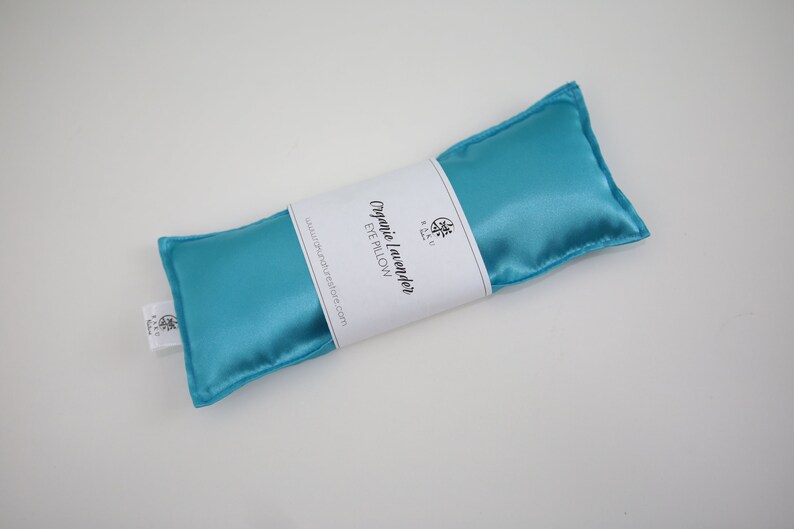 Puede incluir: Una almohada para los ojos de sat&eacute;n azul turquesa con una etiqueta blanca que dice "Organic Lavender Eye Pillow" y una direcci&oacute;n de sitio web.