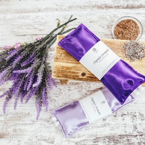 Puede incluir: Dos almohadas para los ojos de sat&eacute;n morado con el texto "Organic Lavender Eye Pillow" en la parte delantera. Las almohadas est&aacute;n sobre una superficie de madera con un ramo de flores de lavanda y un peque&ntilde;o cuenco de lavanda seca.