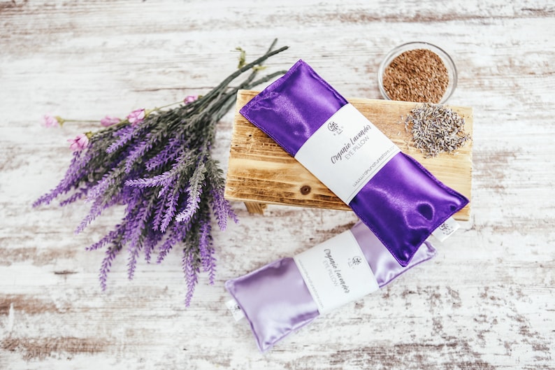 Puede incluir: Dos almohadas para los ojos de sat&eacute;n morado con el texto "Organic Lavender Eye Pillow" en la parte delantera. Las almohadas est&aacute;n sobre una superficie de madera con un ramo de flores de lavanda y un peque&ntilde;o cuenco de lavanda seca.