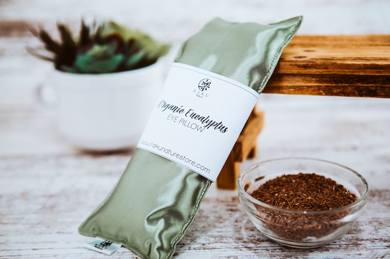 Puede incluir: Una almohada para los ojos de sat&eacute;n verde rellena de eucalipto org&aacute;nico. La almohada tiene una etiqueta blanca que dice "Organic Eucalyptus Eye Pillow" y "www.naturnastore.com".
