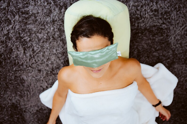 Puede incluir: Una persona con una m&aacute;scara de ojos verde y una toalla blanca, acostada sobre un coj&iacute;n verde en una alfombra gris.
