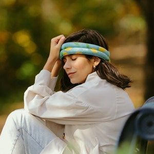 May include: A woman wearing a light blue and yellow head wrap with a lemon pattern. The head wrap is wrapped around the forehead. She is wearing a white shirt and light blue jeans. The background is blurred with green and yellow tones.