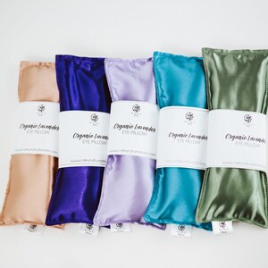 Puede incluir: Cinco almohadas para los ojos de lavanda org&aacute;nica en diferentes colores: melocot&oacute;n, morado, lavanda, azul verdoso y verde. Cada almohada tiene una etiqueta blanca que dice "Organic Lavender Eye Pillow" y una direcci&oacute;n de sitio web.