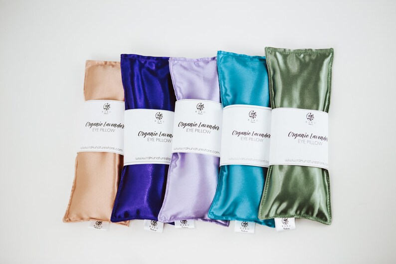 Puede incluir: Cinco almohadas para los ojos de lavanda org&aacute;nica en diferentes colores: melocot&oacute;n, morado, lavanda, azul verdoso y verde. Cada almohada tiene una etiqueta blanca que dice "Organic Lavender Eye Pillow" y una direcci&oacute;n de sitio web.