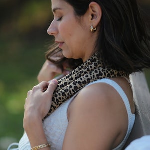 Pode incluir: Uma pessoa usando brincos de argola dourados, uma pulseira de corrente dourada e um lenço com estampa de leopardo. Ela está usando uma regata branca e sentada ao ar livre. O fundo está desfocado com tons de verde e marrom.