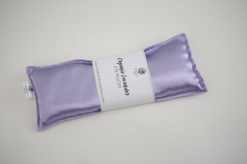 Puede incluir: Una almohada para los ojos de sat&eacute;n lavanda con el texto "Organic Lavender Eye Pillow" y "www.organaturestore.com" impreso en una etiqueta blanca.