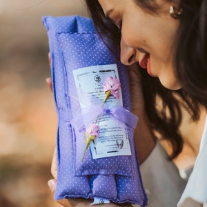 Pode incluir: Uma almofada de aromaterapia com aroma de lavanda em tecido macio, roxo claro, com bolinhas brancas. A almofada é amarrada com uma fita de lavanda e adornada com pequenas flores cor-de-rosa. Uma etiqueta com texto está anexada.