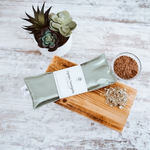 Puede incluir: Una almohada para ojos de color verde con el texto "Organic Eucalyptus Eye Pillow" en la etiqueta. La almohada est&aacute; sobre una superficie de madera con hojas de eucalipto secas y un peque&ntilde;o cuenco de vidrio con hojas de eucalipto.