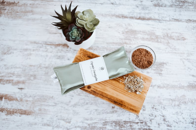 Puede incluir: Una almohada para ojos de color verde con el texto "Organic Eucalyptus Eye Pillow" en la etiqueta. La almohada est&aacute; sobre una superficie de madera con hojas de eucalipto secas y un peque&ntilde;o cuenco de vidrio con hojas de eucalipto.