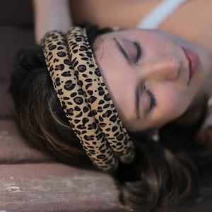 May include: A woman wearing a brown and black leopard print headband with her eyes closed. She is lying on a wooden bench.
