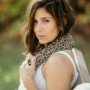 May include: A woman with brown hair wearing a white tank top and a leopard print scarf. She is wearing a gold chain bracelet and blue jeans.