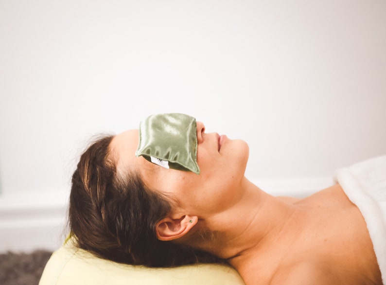 Puede incluir: Una persona acostada de espaldas con una almohadilla para los ojos verde que le cubre los ojos. La persona lleva una toalla blanca y tiene el pelo recogido hacia atr&aacute;s.