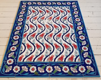 Op de afbeelding: Rechthoekige tegels met een centraal patroon van rode tulpen en blauwe stelen op een witte achtergrond. De rand is een blauw bloemmotief met witte bloemen. De tegels liggen op een witte houten vloer.