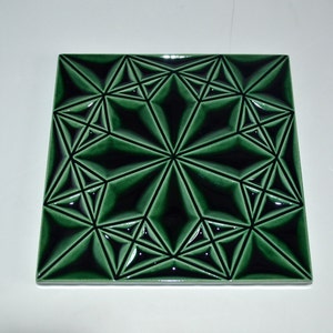 Op de afbeelding: Vierkante, smaragdgroene tegel met een geometrisch sterpatroon. Het ontwerp heeft een reeks onderling verbonden ruit- en driehoekvormen, waardoor een driedimensionaal effect ontstaat. De tegel heeft een glanzende afwerking, ongeveer 20 cm x 20 cm.