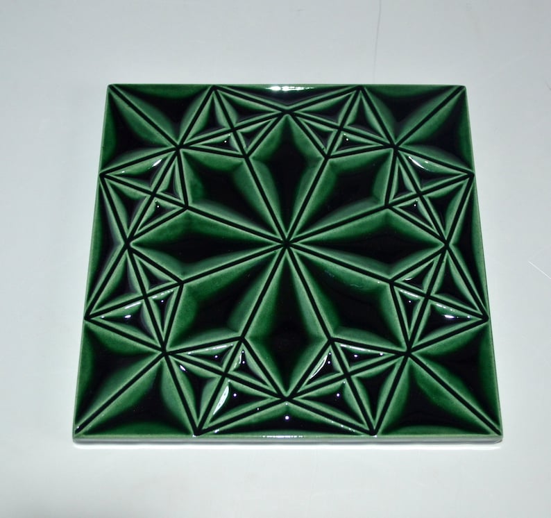Op de afbeelding: Vierkante, smaragdgroene tegel met een geometrisch sterpatroon. Het ontwerp heeft een reeks onderling verbonden ruit- en driehoekvormen, waardoor een driedimensionaal effect ontstaat. De tegel heeft een glanzende afwerking, ongeveer 20 cm x 20 cm.