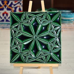 Puede incluir: Azulejo cerámico verde esmeralda cuadrado con un patrón de estrella geométrico en relieve. El azulejo se exhibe sobre un pequeño caballete de madera. El diseño presenta un patrón repetitivo de formas de estrella, creando una superficie texturizada. Otros azulejos son visibles en el fondo.