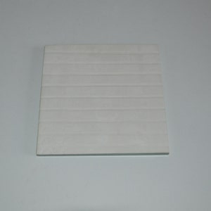 Op de afbeelding: Een vierkante, gebroken witte onderzetter met een gestructureerd, horizontaal gestreept patroon. De onderzetter is gemaakt van een stevig materiaal en is ontworpen om oppervlakken te beschermen tegen condensatie van drankjes. De onderzetter heeft een neutrale kleur.