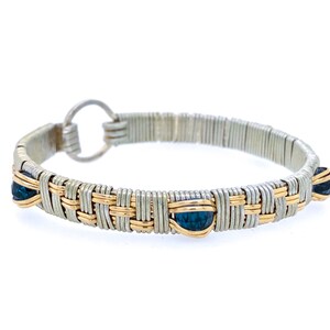May include: A silver and gold-toned wire-wrapped bracelet with three dark blue gemstones. The bracelet has a woven design and a circular clasp. The gemstones are teardrop-shaped.