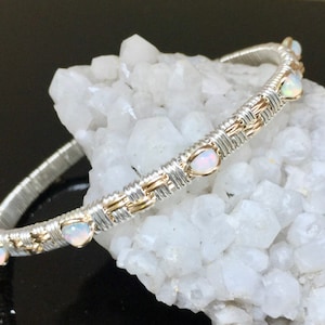 Könnte beinhalten: Ein silber- und goldfarbenes, drahtgewickeltes Armband mit fünf weißen Opal-Steinen. Das Armband liegt auf einem weißen Kristall-Hintergrund.