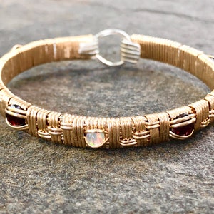 Könnte beinhalten: Goldfarbenes, drahtgewickeltes Armband mit gewebtem Design. Das Armband hat abwechselnd rote und irisierende Edelsteine. Der Verschluss ist silberfarben. Ein handgefertigtes Schmuckstück.