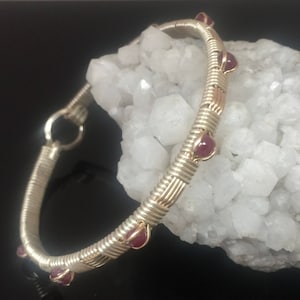 May include: A silver-coloured bangle bracelet with a woven design, featuring small, round, red gemstones. The bracelet is displayed on a white, crystalline surface, with a dark background.