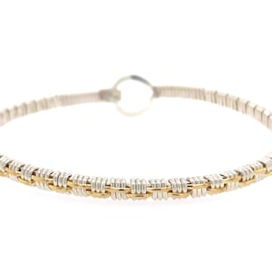 May include: A silver and gold-toned wire-wrapped bangle bracelet. The bracelet features a woven design with alternating sections of silver and gold-colored wire. The bracelet has a circular shape and is designed to be worn on the wrist.