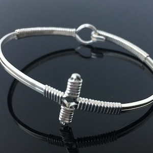 May include: A silver bracelet featuring a cross design. The cross is centrally placed and has a wrapped wire pattern. The bracelet has a hook and eye clasp. It is displayed on a reflective black surface.
