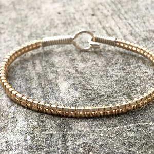 May include: Gold-coloured wire-wrapped bracelet with a hook clasp. The bracelet has small, square-shaped gold beads, creating a textured look. The clasp and connecting wire are silver-coloured.