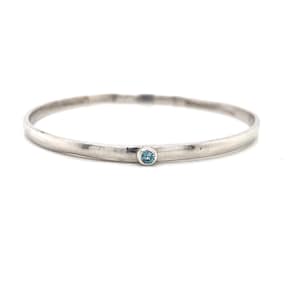 Può includere: Un braccialetto rigido in argento con una singola pietra preziosa blu incastonata al centro.