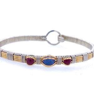 Può includere: Un braccialetto avvolto in filo d'argento e oro con tre accenti di pietre preziose. Il braccialetto presenta una gemma blu ovale centrale affiancata da due gemme rosse rotonde più piccole. La chiusura è una semplice chiusura a gancio e occhiello.