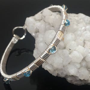 May include: A silver and gold wire-wrapped bangle bracelet with teardrop-shaped, light blue gemstones. The bracelet is displayed on a white, crystalline surface against a dark background. The bracelet's design incorporates silver and gold wire.