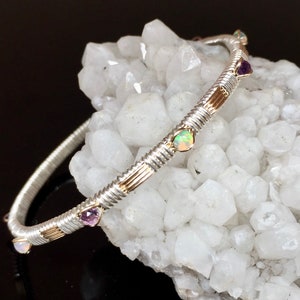 May include: A silver and gold bangle bracelet with opal and amethyst gemstones. The bracelet has a twisted wire design with gold accents, set against a white crystal background. The gemstones are round and oval shaped.
