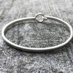 May include: A silver bangle bracelet with a ribbed texture and a hook clasp. The bracelet is displayed on a grey, textured surface. The bracelet's design is simple and elegant.
