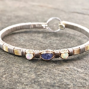 May include: Silver and gold wire-wrapped bracelet with three gemstones. The bracelet features a central blue gemstone flanked by two white gemstones. The clasp is a simple hook and eye closure. The bracelet is displayed on a grey surface.